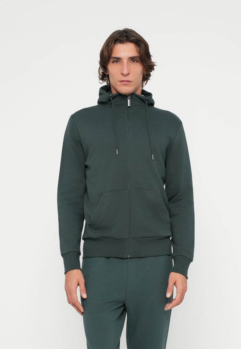 Kapuzenpullover mit Reißverschluss in dunkelgrünem Baumwollmischgewebe, ausgestattet mit zwei vorderen Taschen, Kapuze mit Kordelzug sowie gerippten Bündchen und Saum. Durchgehend glatte Textur.