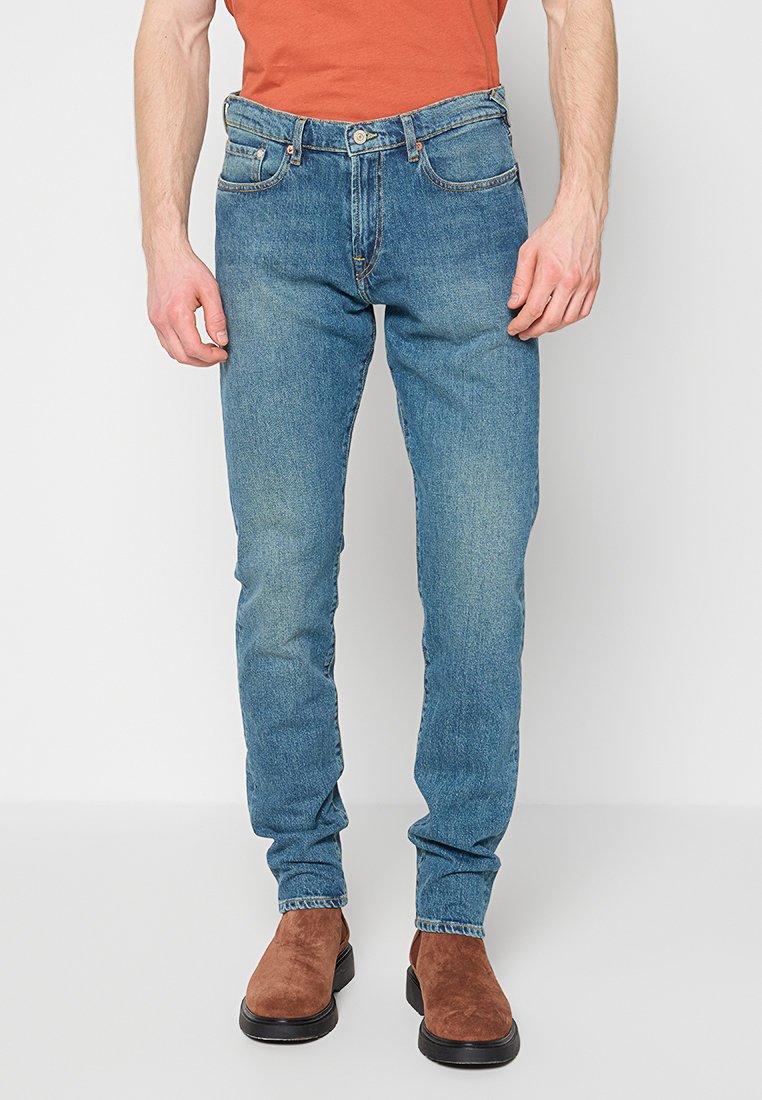 Blaue Denim-Jeans mit schlanker Passform, ausgestattet mit fünf Taschen, einem Knopfverschluss und leichtem Used-Look. Kombiniert mit braunen Stiefeletten.