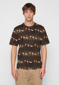 Mann trägt ein dunkelbraunes T-Shirt mit wiederholtem Aufdruck von Sonnenbrillen und Eiskaffeebecher, kombiniert mit beigen Hosen, steht vor einem grauen Hintergrund.