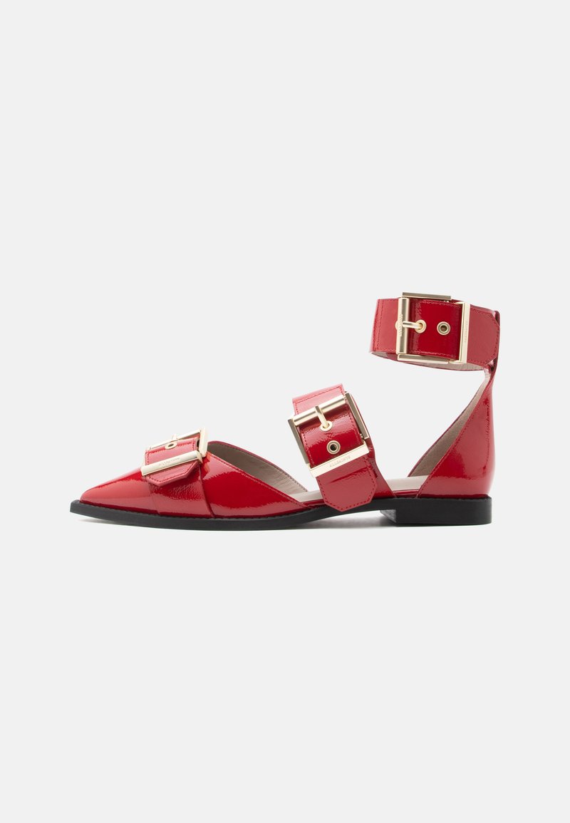 Roter flacher Schuh aus Lackleder mit spitzer Kappe, drei großen goldenen Schnallen über Fuß und Knöchel sowie schwarzer Sohle auf weißem Hintergrund.