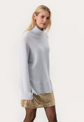 Blonde Frau trägt einen hellgrauen, übergroßen Rollkragenpullover, einen goldenen Paillettenminirock, schwarze Strumpfhosen und goldene Creolen.