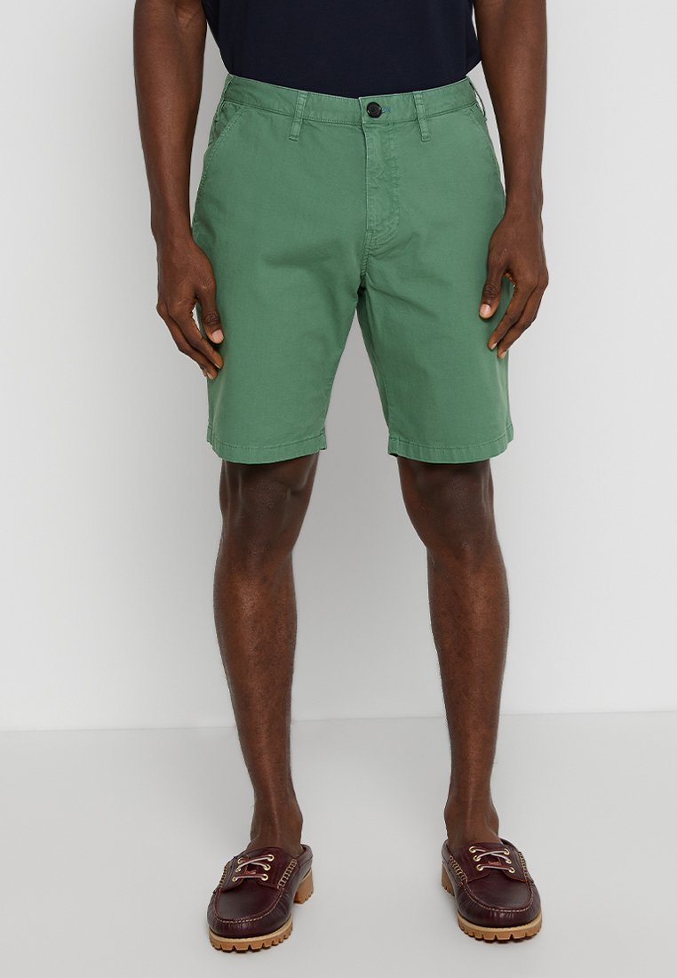 Grüne Baumwollshorts mit Knopfverschluss, Fronttaschen und leicht umgeschlagenen Säumen; kombiniert mit dunkelbraunen Schuhen.