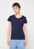 Person mit kurzen braunen Haaren, die ein marineblaues T-Shirt mit V-Ausschnitt, kleinem Logo und hellblauen Jeans vor einem einfarbigen Hintergrund trägt.