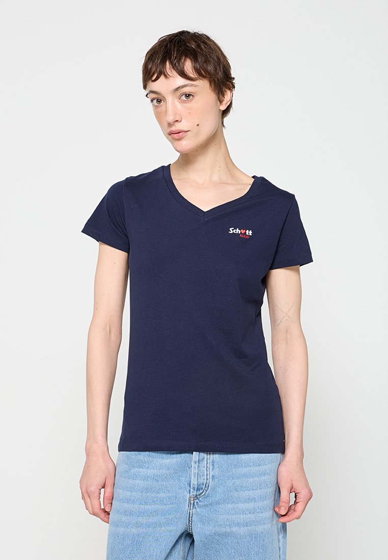 Person mit kurzen braunen Haaren, die ein marineblaues T-Shirt mit V-Ausschnitt, kleinem Logo und hellblauen Jeans vor einem einfarbigen Hintergrund trägt.