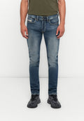 Mann in schmal geschnittenen, ausgewaschenen blauen Jeans, schwarzen und grauen Sneakers sowie einem olivgrünen T-Shirt, der vor einem schlichten weißen Hintergrund steht.