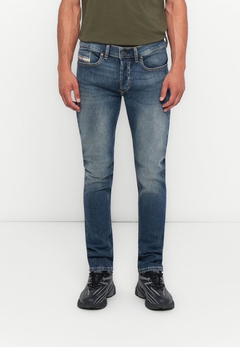 Mann in schmal geschnittenen, ausgewaschenen blauen Jeans, schwarzen und grauen Sneakers sowie einem olivgrünen T-Shirt, der vor einem schlichten weißen Hintergrund steht.