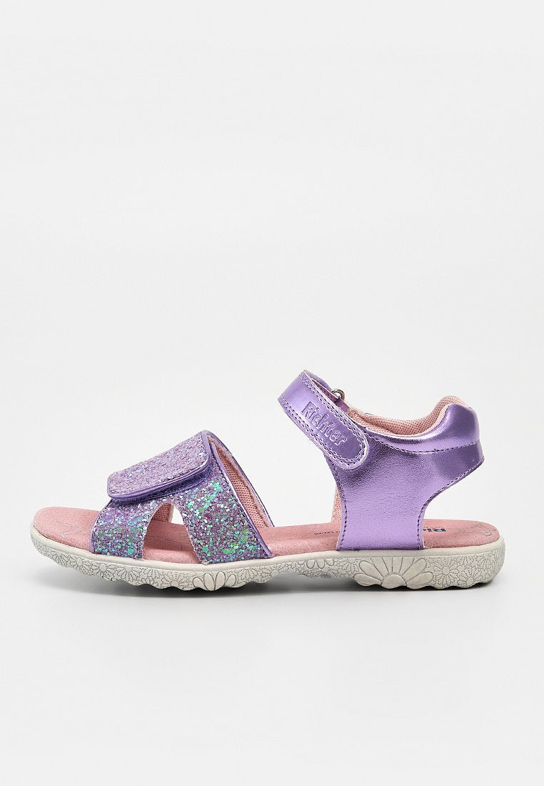 Lila und glitzernde Kindersandale mit zwei verstellbaren Riemen und einer pinken gepolsterten Innensohle auf einer weißen, mit Blumenmuster versehenen Sohle.