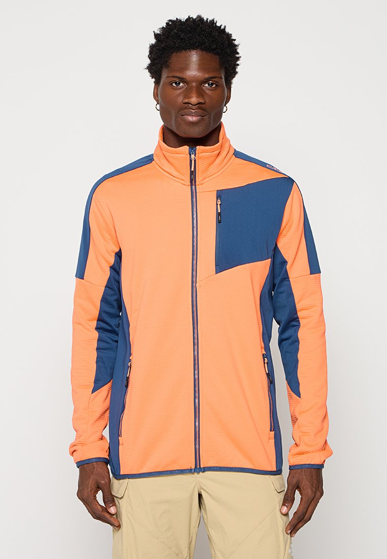 Mann in einer orange- und dunkelblauen Jacke mit Reißverschlüssen und Reißverschlusstaschen, steht vor einem schlichten hellen Hintergrund.