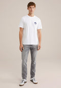 Junger Mann steht, trägt ein weißes Grafik-T-Shirt, graue Jeans und weiße Sneakers mit schwarzen Streifen vor einem schlichten beigen Hintergrund.