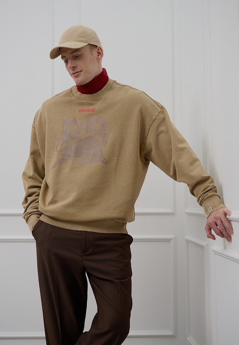 Tailliertes Sweatshirt mit Rippstruktur, rotem "HUGO"-Schriftzug und Grafik, getragen über einem roten Rollkragenpullover, kombiniert mit braunen Hosen und passender Kappe.
