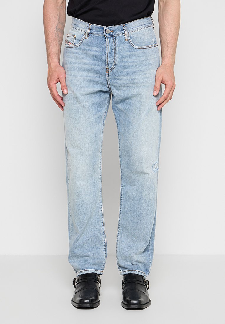 Mann trägt hellblaue Straight-Leg-Jeans mit leichter Abnutzung am Knie, schwarze Lederschnallenschuhe und ein schwarzes, eingestecktes Hemd.