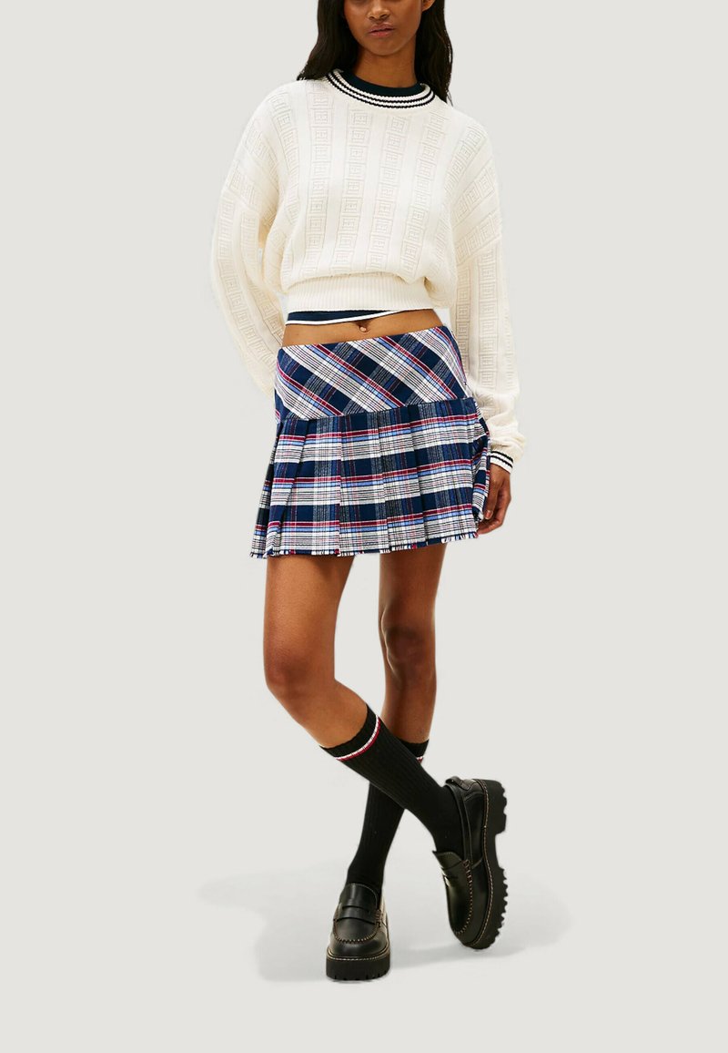 Weißer Strickpullover mit strukturiertem Muster, kombiniert mit einem blauen karierten Rock mit roten und weißen Akzenten. Schwarze knielange Socken und Schuhe.