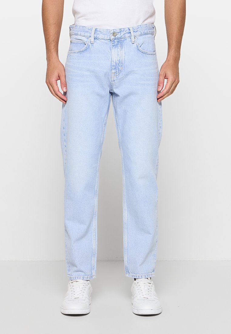 Mann trägt hellblaue, gerade geschnittene Jeans, weiße Sneakers und ein weißes Hemd und steht vor einem einfarbigen Hintergrund.