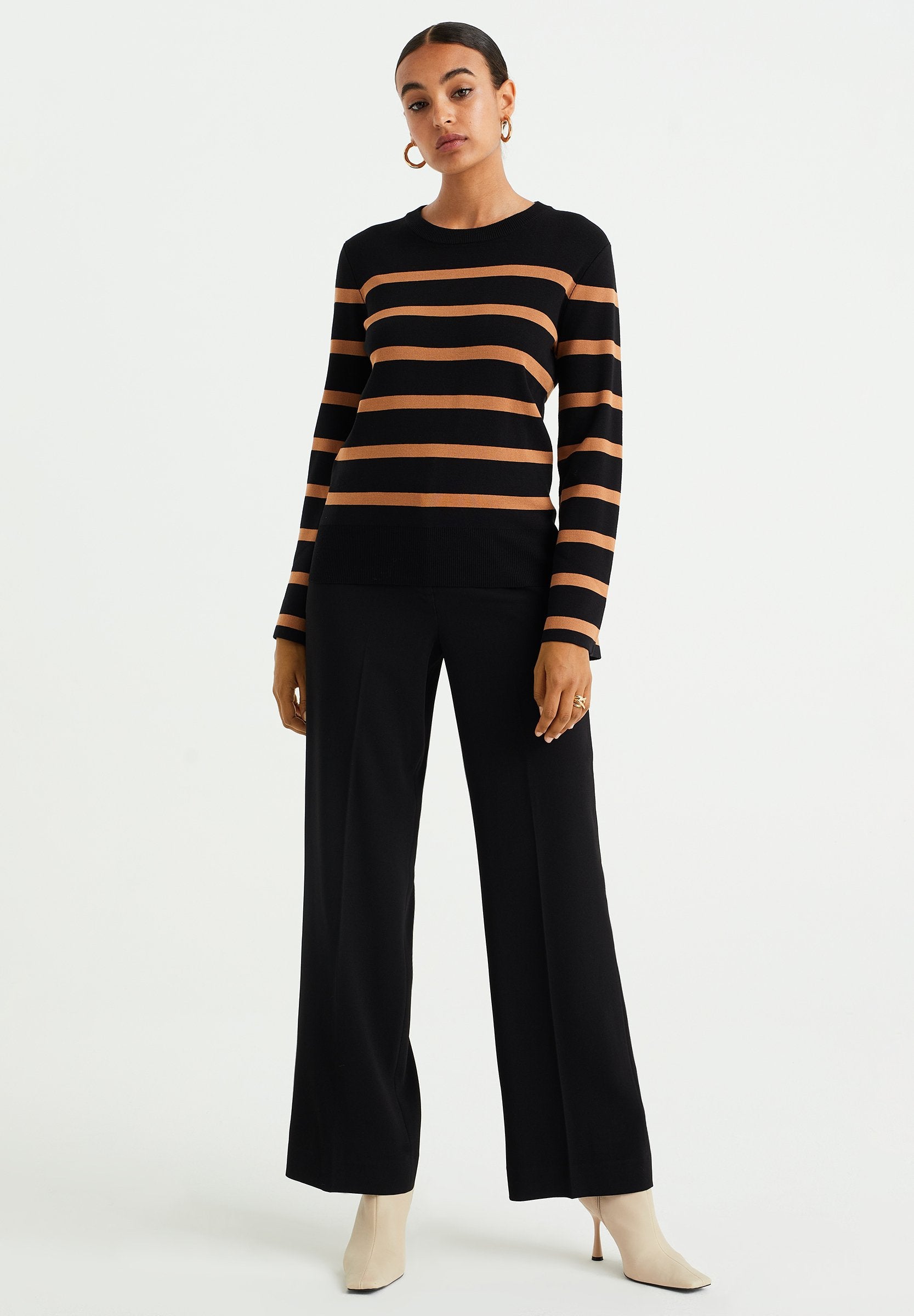 Frau steht und trägt ein gestreiftes Langarmoberteil in Schwarz und Braun, schwarze weite Hosen, beige High-Heel-Stiefeletten und Creolen.
