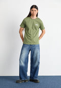 Olivgrünes T-Shirt mit kurzen Ärmeln, lockerer Schnitt, mit weißem Logo. Kombiniert mit weit geschnittenen blauen Jeans und schwarzen Plateaustiefeln. Minimalistisches Design.