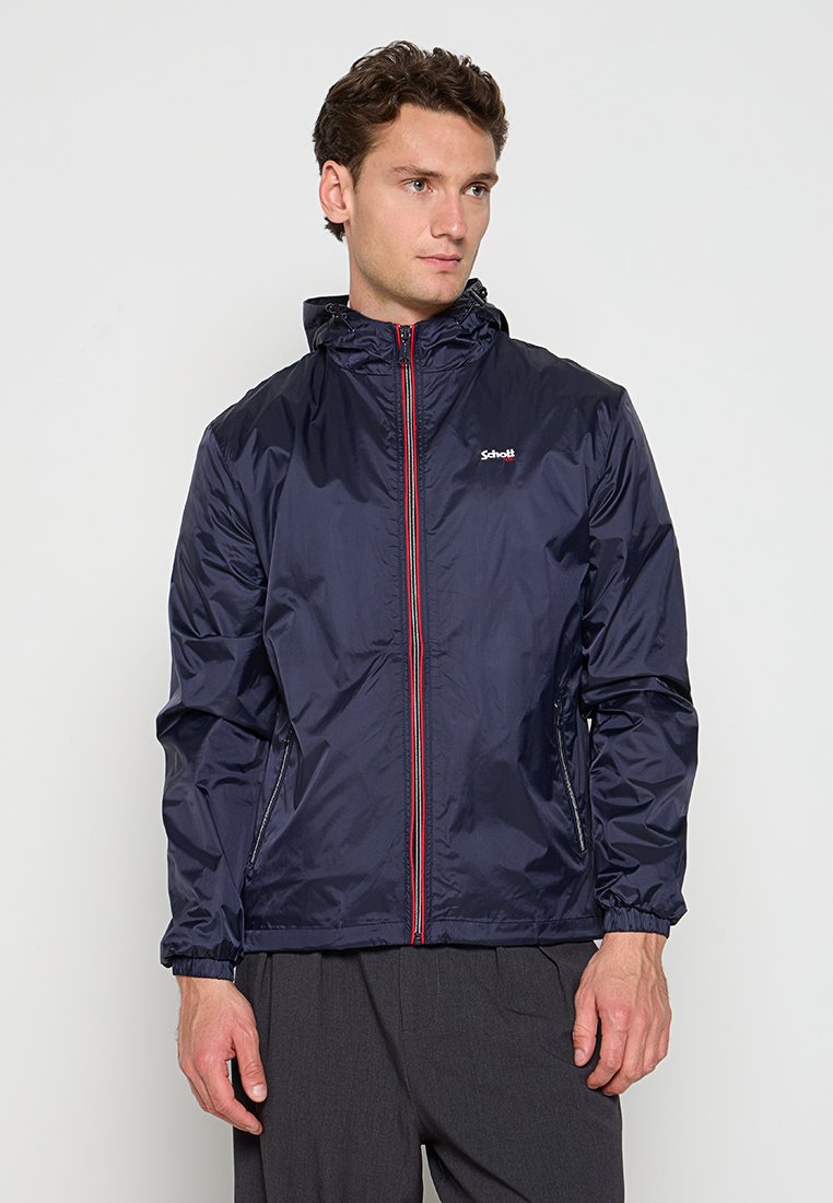 Marineblauer Windbreaker mit Kapuze, ausgestattet mit einem roten Reißverschluss und elastischen Bündchen. Hergestellt aus glattem, leichtem Material. Dezentes Logo auf der Brust.