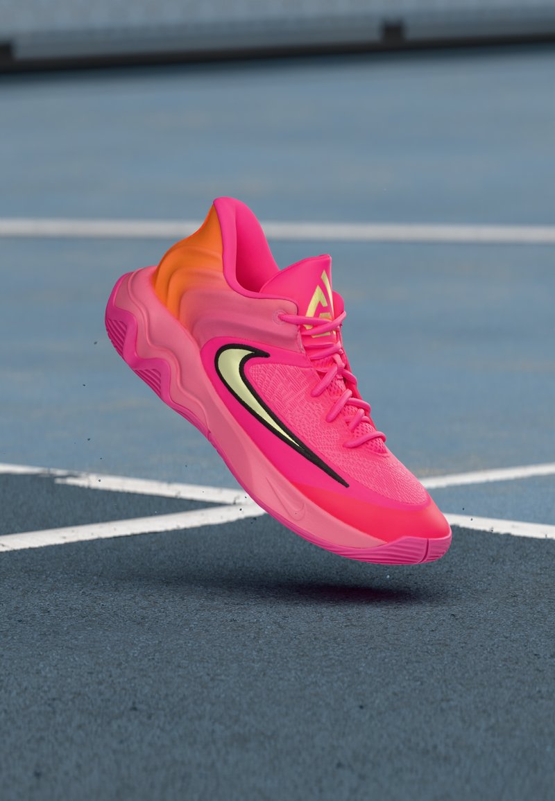 Rosa Sportschuhe mit einem farblichen Verlauf zum orangefarbenen Absatz, strukturiertem Obermaterial, schwarzem Akzentstreifen und sichtbaren Schnürsenkeln, erhöht über einer blauen Fläche.