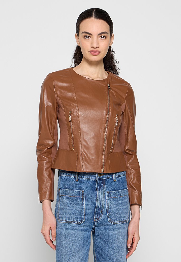 Frau trägt eine taillierte braune Lederjacke mit asymmetrischen Frontreißverschlüssen und blaue hochgeschnittene Jeans, stehend vor einfachem Hintergrund.