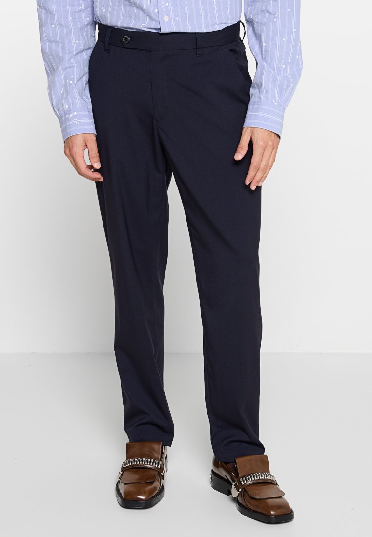Marineblaue Hose mit taillierter Passform, aus glattem Stoff, mit Gürtelschlaufen und Seitentaschen. Kombiniert mit einem hellen, gemusterten Hemd und braunen Schuhen.