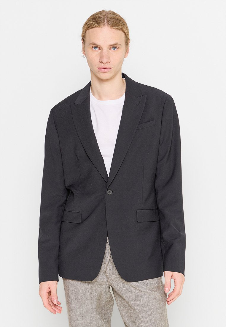 Schwarzes Blazer mit eingekerbten Revers, einem Knopfverschluss und zwei vorderen Taschen, getragen über einem weißen T-Shirt. Glattes, strukturiertes Material.