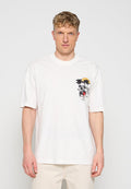 Mann mit lockigem Haar, der ein weißes T-Shirt mit kleinem Totenkopf, Palmen und Sonne auf der linken Brust trägt, steht vor hellem Hintergrund.