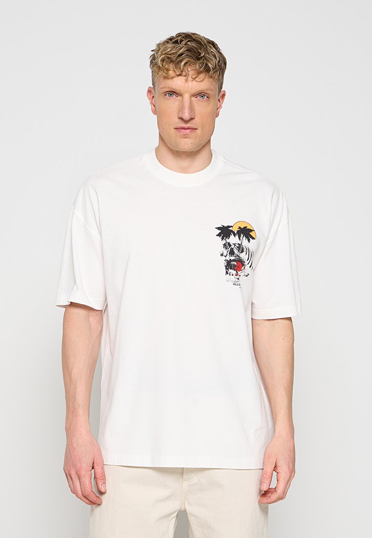 Mann mit lockigem Haar, der ein weißes T-Shirt mit kleinem Totenkopf, Palmen und Sonne auf der linken Brust trägt, steht vor hellem Hintergrund.