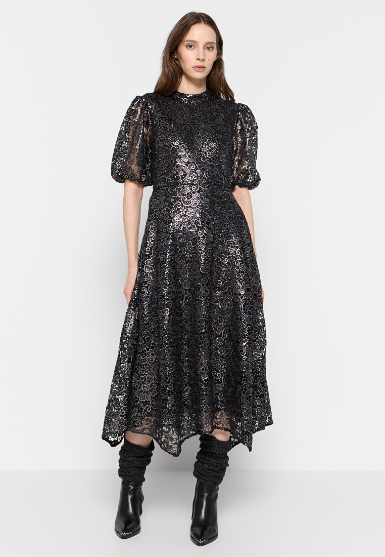 Schwarzes Spitzenkleid mit Puffärmeln, das florale Muster aufweist. Knielang mit fließendem Rock. Das Model kombiniert es mit schwarzen Knöchelschuhen und strukturierten Beinstulpen.
