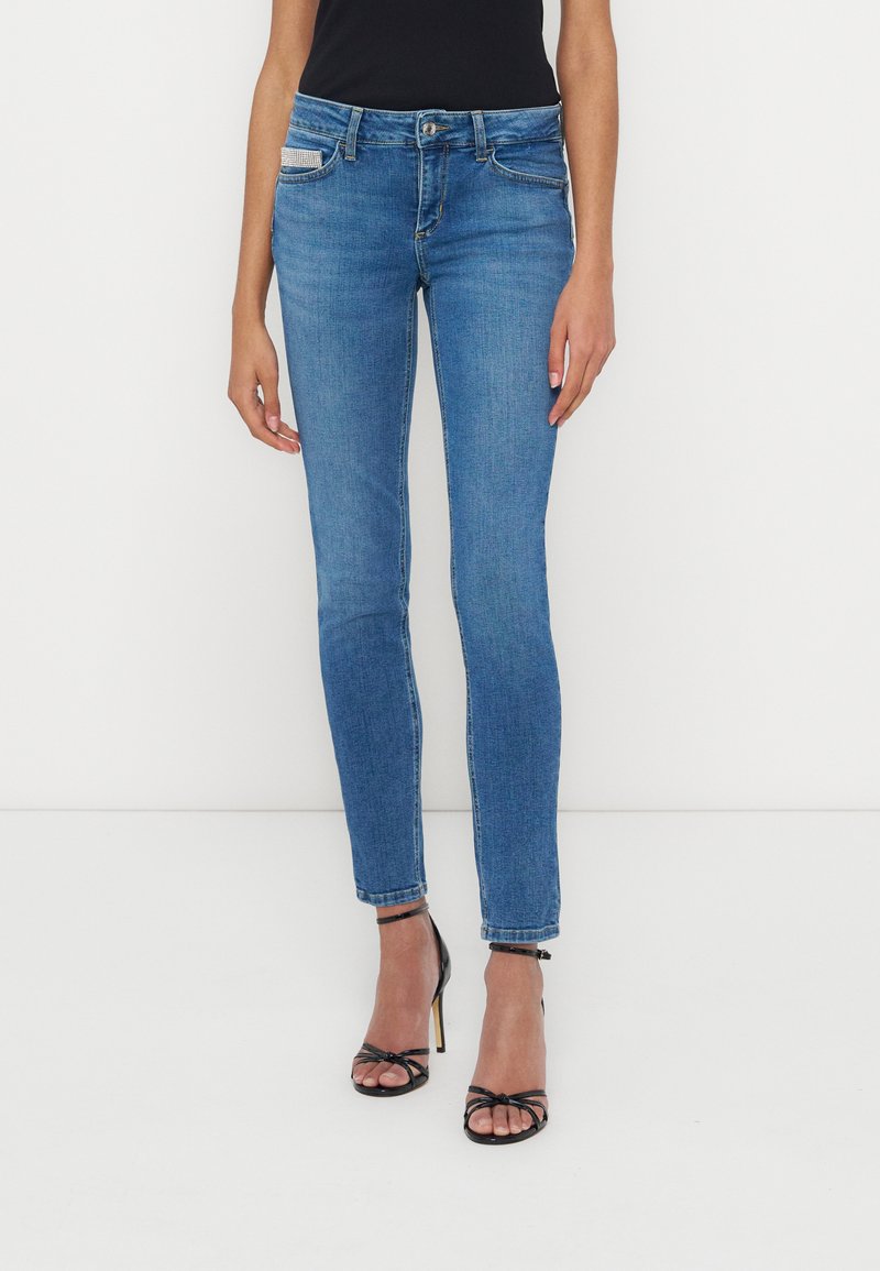 Frau trägt eine eng anliegende blaue Jeans und schwarze Riemchen-High-Heel-Sandalen und steht vor einem weißen Hintergrund.