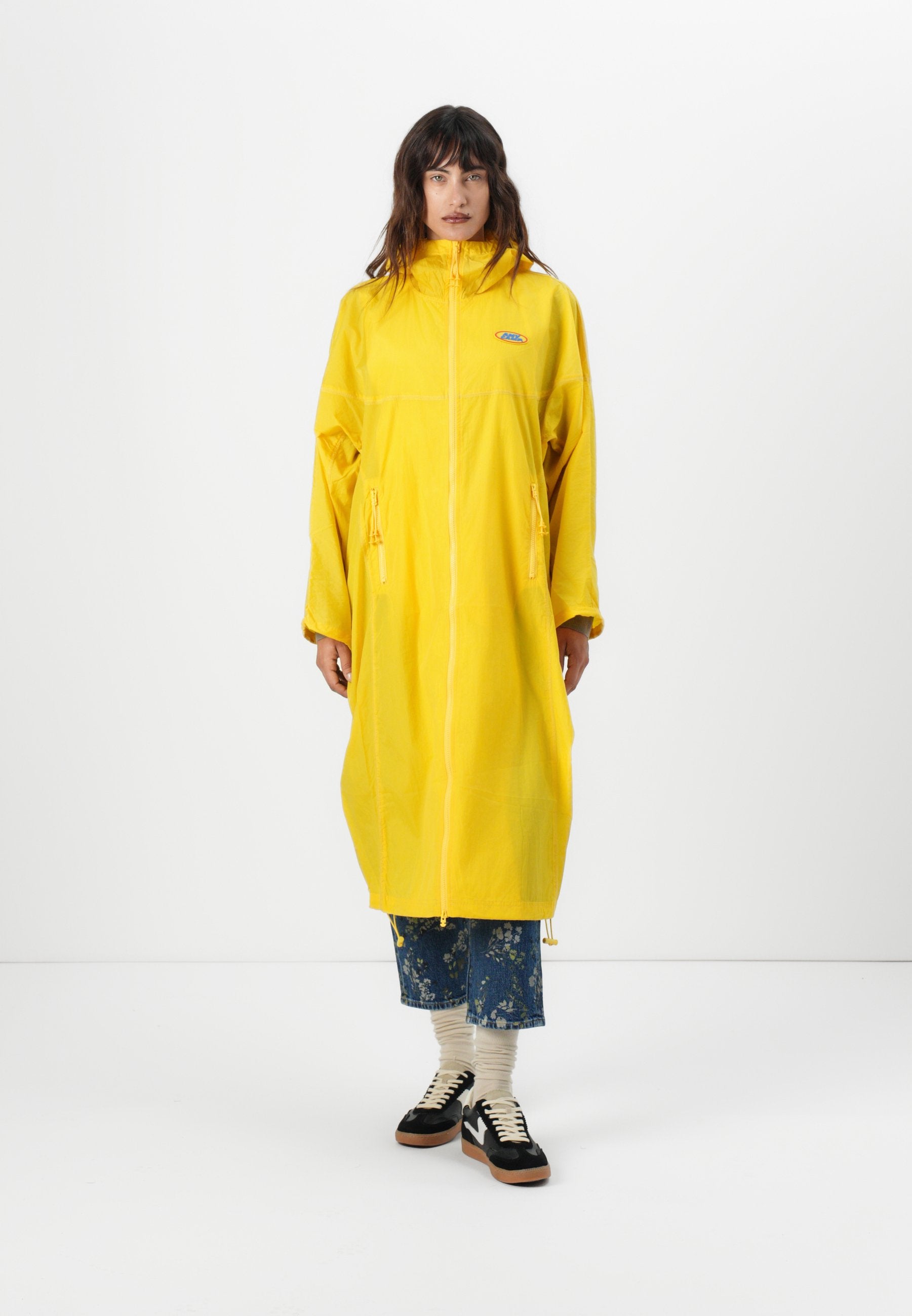 Gelber, langer Regenmantel mit vollem Reißverschluss, hohem Kragen, elastischen Bündchen und Fronttaschen. Getragen über einer blauen Hose mit Blumenmuster und schwarzen Sneakers.