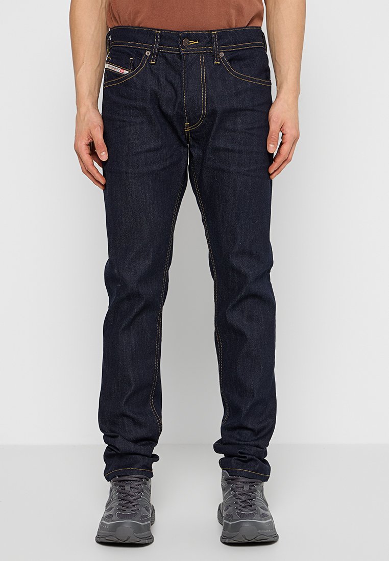 Dunkelblaue Jeans mit geradem Schnitt, gelben Nähten, fünf Taschen und einem Knopfverschluss. Getragen mit grauen Turnschuhen.