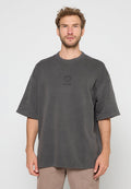 Mann mit heller Haut und lockigem Haar trägt ein übergroßes, dunkelgraues T-Shirt mit kleinem Logo und steht vor einem schlichten weißen Hintergrund.