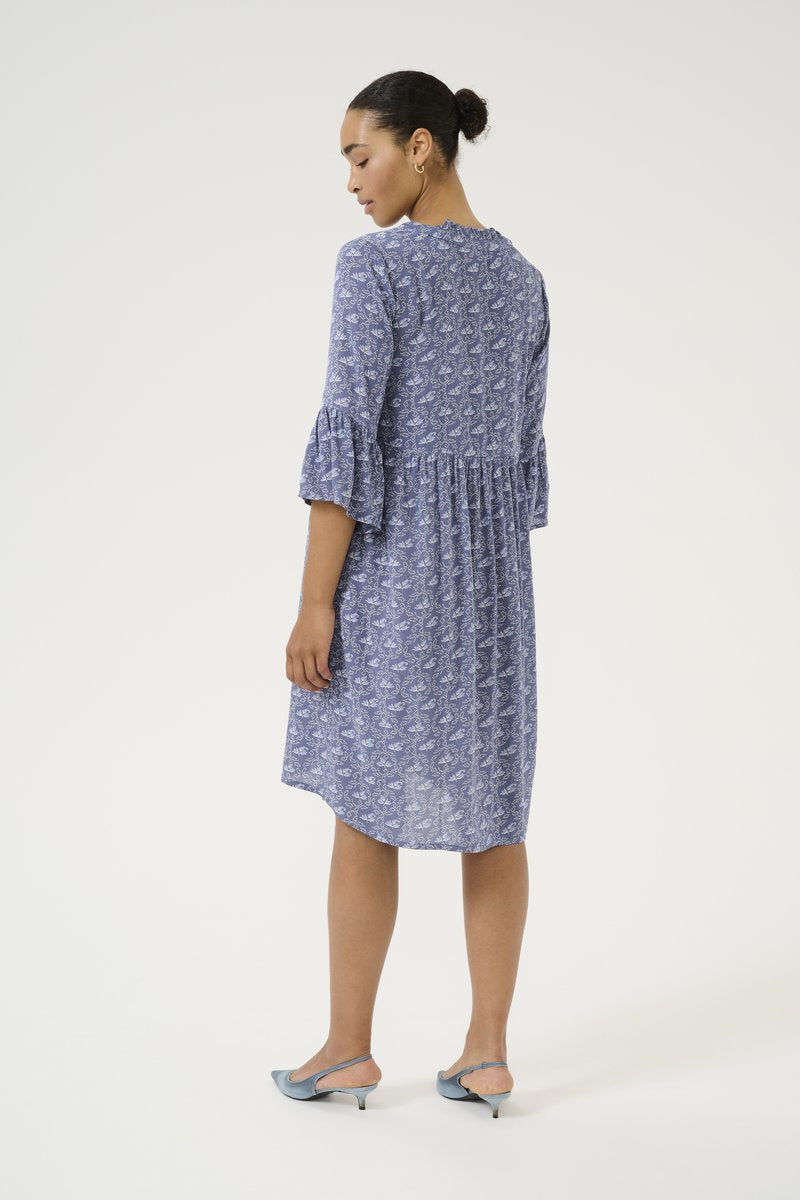 Blau gemustertes Kleid mit Rüschensleeves und geraffter Taille. Runder Ausschnitt. Getragen mit blauen Slingback-Absätzen; glatte Stoffbeschaffenheit.