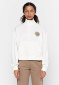 Frau mit lockigem Haar, die einen weißen Sweatshirt mit Viertelreißverschluss und einem bunten runden Logo sowie beigen Hosen mit Text auf dem Oberschenkel trägt.