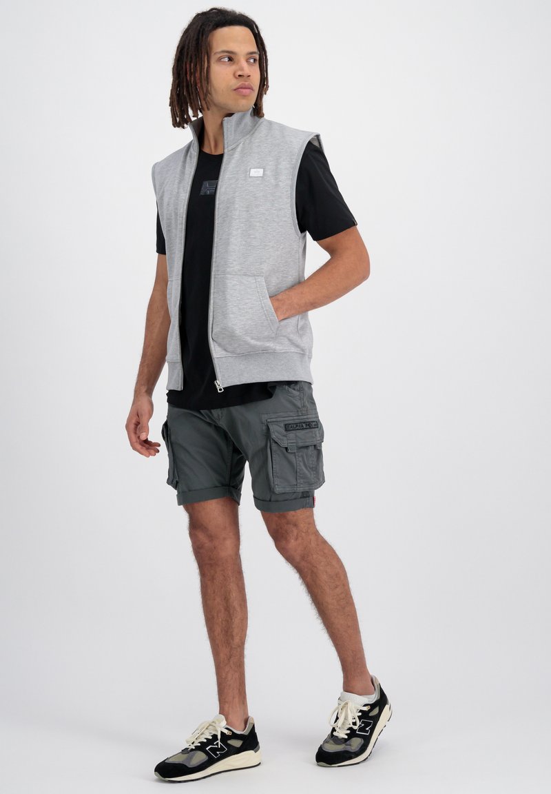 Graue ärmellose Zip-Weste über einem schwarzen T-Shirt, kombiniert mit dunkelgrauen Cargo-Shorts und schwarzen Sneakers mit weißen Akzenten und Taschen.