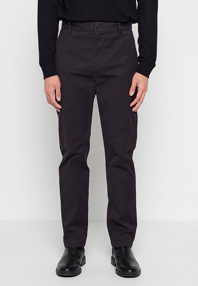 Mann in schwarzen Slim-Fit-Hosen, schwarzem Sweatshirt und schwarzen Lederstiefeln, der vor einem schlichten weißen Hintergrund steht.