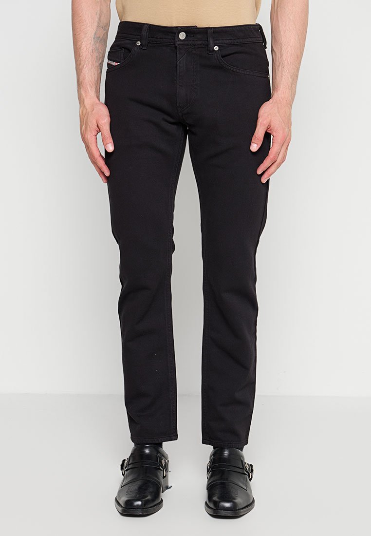 Mann trägt eng anliegende schwarze Jeans und schwarze Leder-Monk-Strap-Schuhe, steht vor einem schlichten weißen Hintergrund.