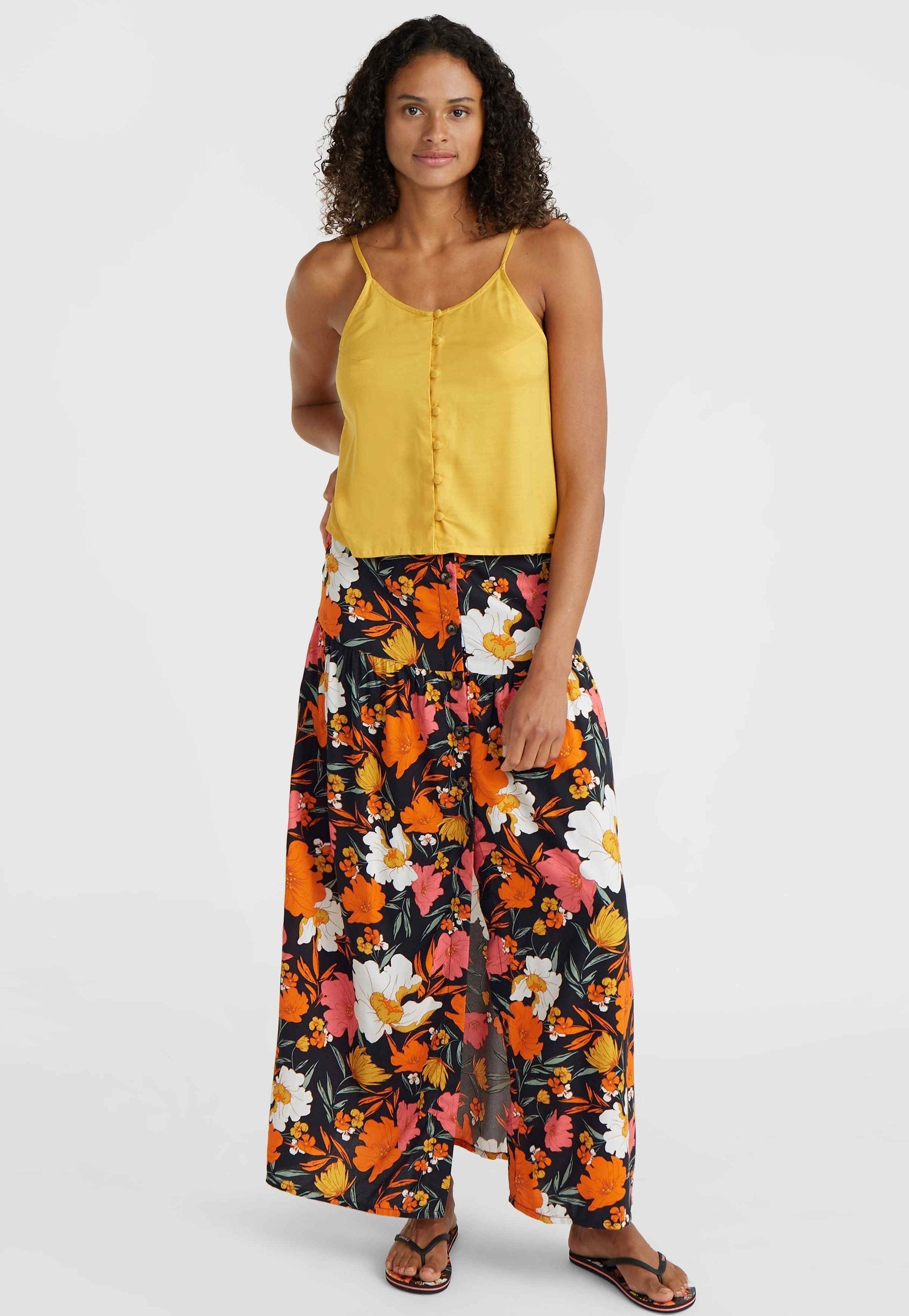 Gelbe Satin-Camisole mit Spaghettiträgern und Knopfdetails, kombiniert mit einem luftigen schwarzen Rock mit bunten floralen Mustern.