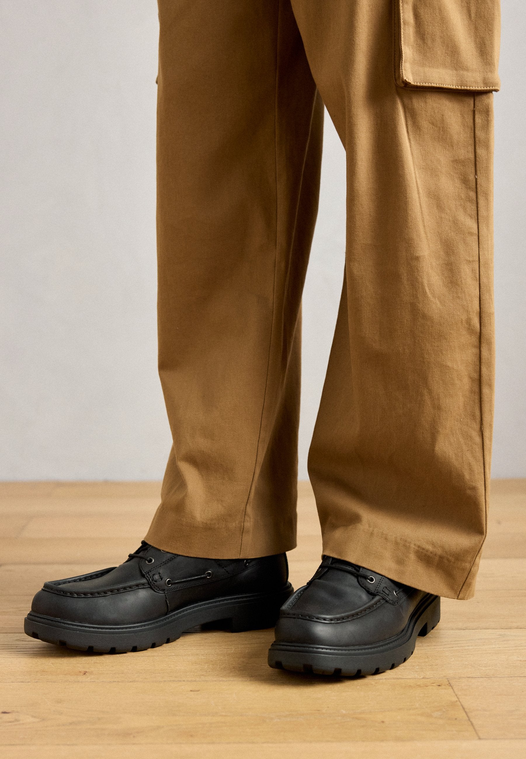 Schwarze Lederschuhe mit einer massiven Gummisohle, detaillierten Nähten und Schnürakzenten. Kombiniert mit hellbraunen, weit geschnittenen Hosen auf einem Holzboden.
