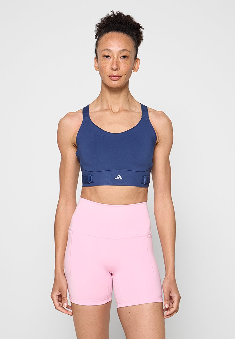 Fit Frau mit lockigem Haar trägt dunkelblauen Sport-BH und hoch taillierte pinke Radlerhose, steht vor hellem einfarbigem Hintergrund.