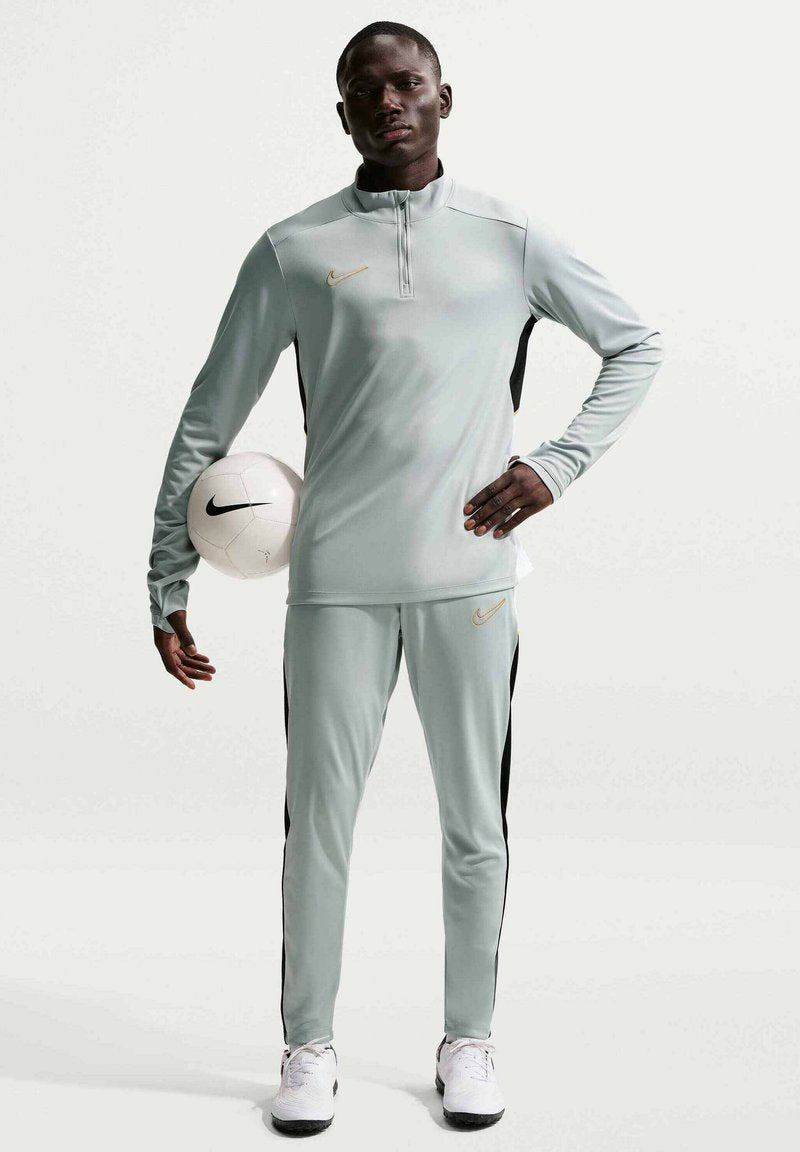 Mann in grauem Nike-Trainingsanzug hält mit einer Hand einen weißen Nike-Fußball und steht vor einem schlichten Hintergrund.