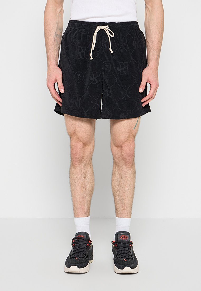 Mann trägt schwarze Shorts mit Kordelzug, weiße Socken und schwarze Sneaker, steht vor einfarbigem Hintergrund.