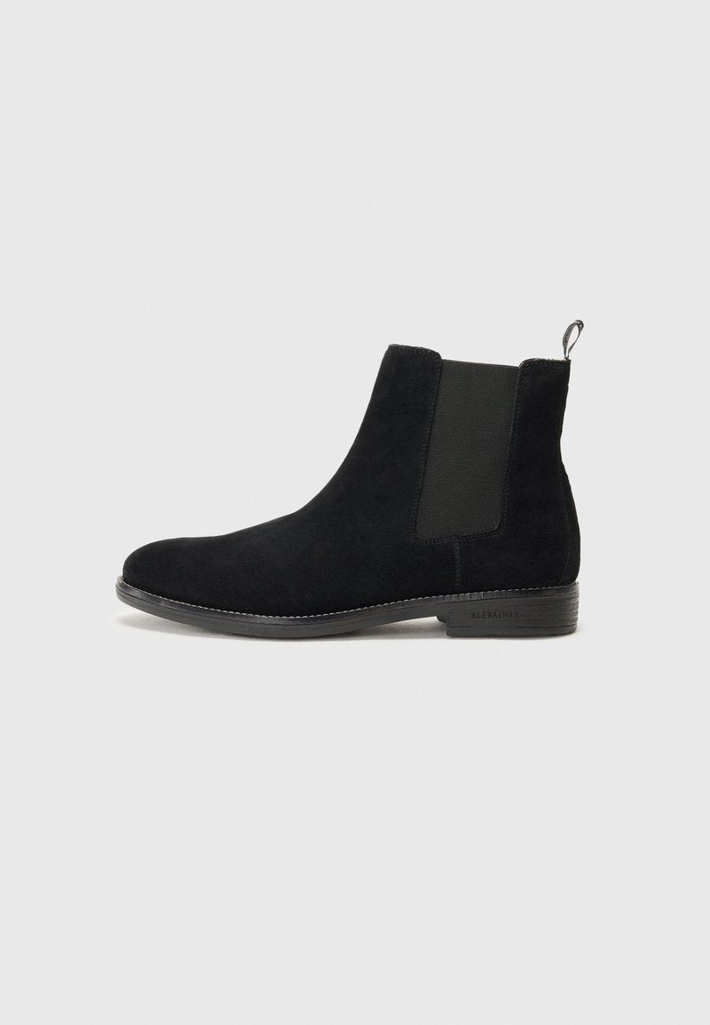 Schwarze Wildleder Chelsea-Boots mit elastischen Seiteneinsätzen und einer Zuglasche an der Ferse. Flache Sohle mit dezentem Branding an der Seite.
