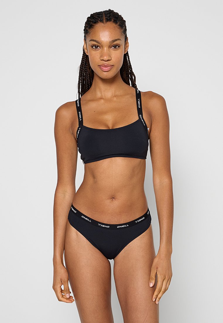 Frau mit geflochtenem Haar trägt einen schwarzen Bikini mit "O'Neill" gebrandeten Trägern und Bund, sie steht vor einem einfarbigen hellen Hintergrund.