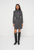 Schwarzes Kleid mit weißen Polka-Dots, langen Ärmeln und hohem Kragen, in der Taille mit einem Gürtel betont, kombiniert mit hohen schwarzen Stiefeln mit Absatz.