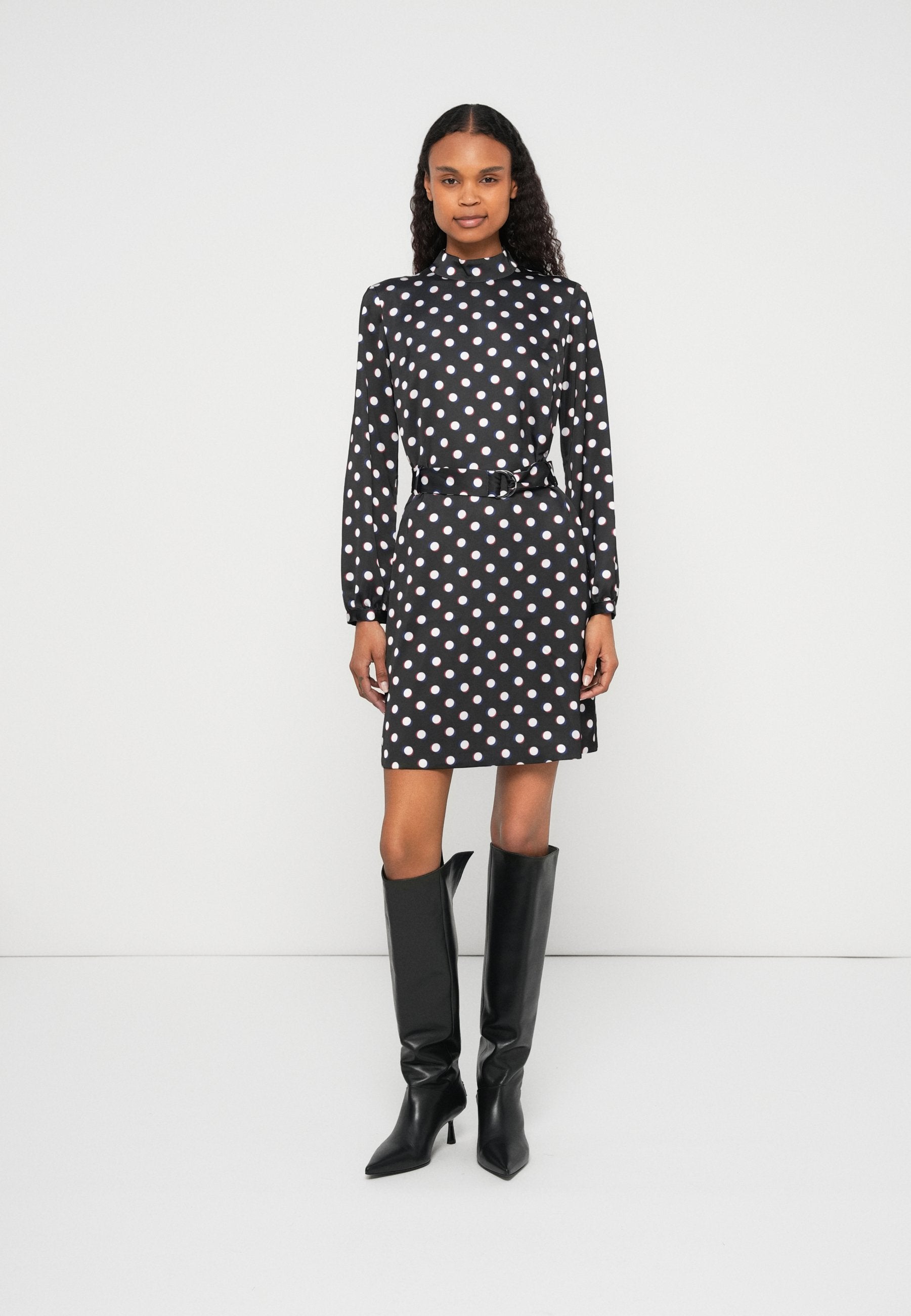 Schwarzes Kleid mit weißen Polka-Dots, langen Ärmeln und hohem Kragen, in der Taille mit einem Gürtel betont, kombiniert mit hohen schwarzen Stiefeln mit Absatz.