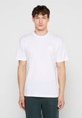 Junger Mann, der nach vorne blickend steht, trägt ein einfaches weißes T-Shirt mit kurzen Ärmeln und dunkle Hosen vor einem hellgrauen Hintergrund.