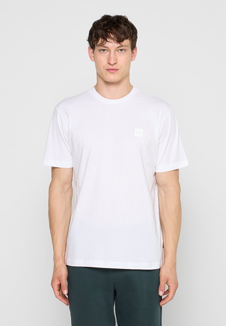 Junger Mann, der nach vorne blickend steht, trägt ein einfaches weißes T-Shirt mit kurzen Ärmeln und dunkle Hosen vor einem hellgrauen Hintergrund.