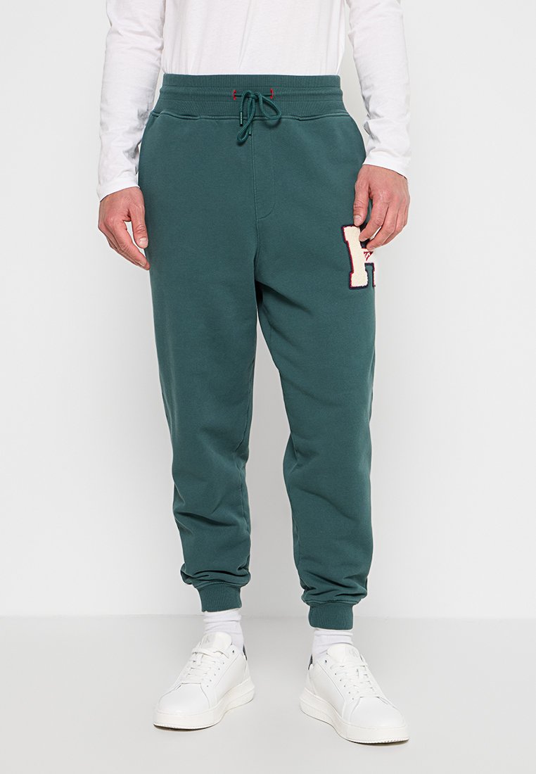 Mann trägt türkisfarbene Jogginghosen mit verstellbarem Bund, weiße Socken und weiße Sneakers und steht vor einem schlichten Hintergrund.