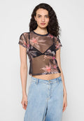 Frau mit lockigem dunklem Haar trägt einen schwarzen, transparenten, floralen Top über einem schwarzen BH und hoch taillierte hellblaue Jeans, stehend vor einem schlichten Hintergrund.