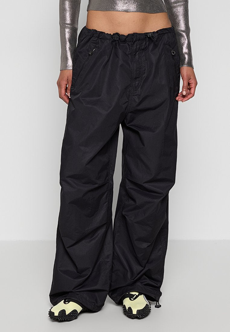 Person in lockeren schwarzen Reißverschluss-Hosen, metallischem, langärmligem Cropped-Top und creme- und schwarzfarbigen, strukturierten Sneakers steht vor einem schlichten Hintergrund.